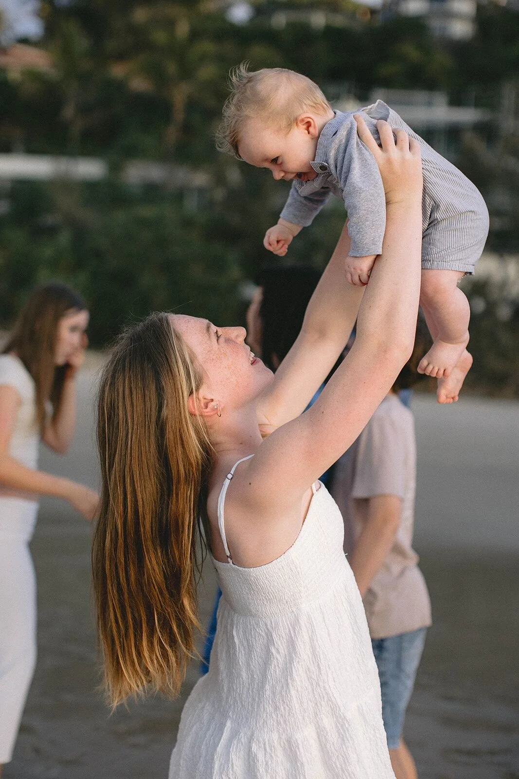 Teenage girl lifting a laughing baby boy into the air on the beach