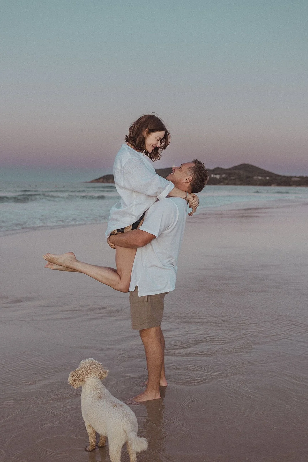 Man lifting his fiancée in the shallows with the headland and their dog behind, Jamie and Jessica's beach marriage proposal at Belongil Beach, Byron Bay