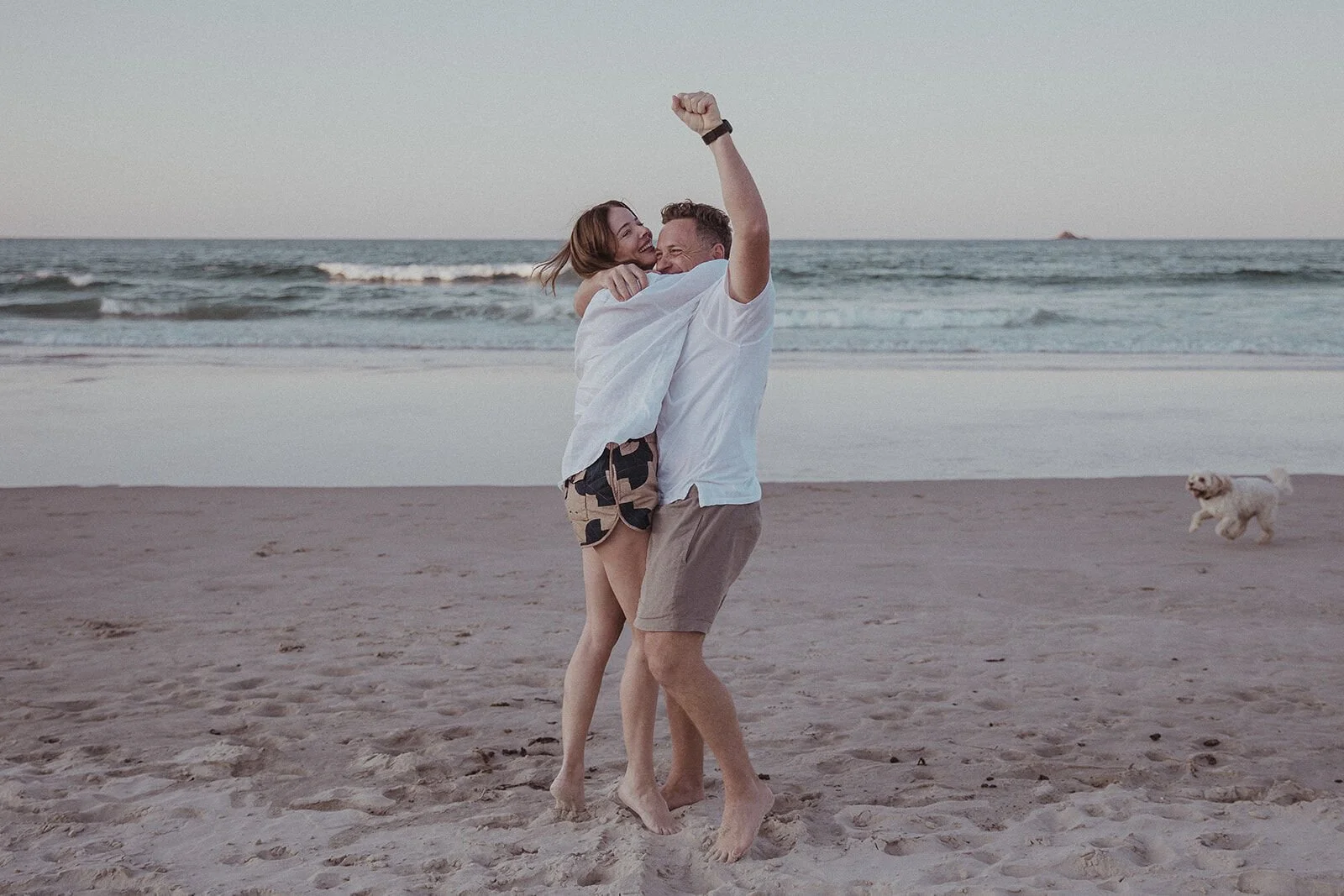 Man lifting the woman, fist in the air, as their dog runs across the sand, Jamie and Jessica's beach marriage proposal at Belongil Beach, Byron Bay