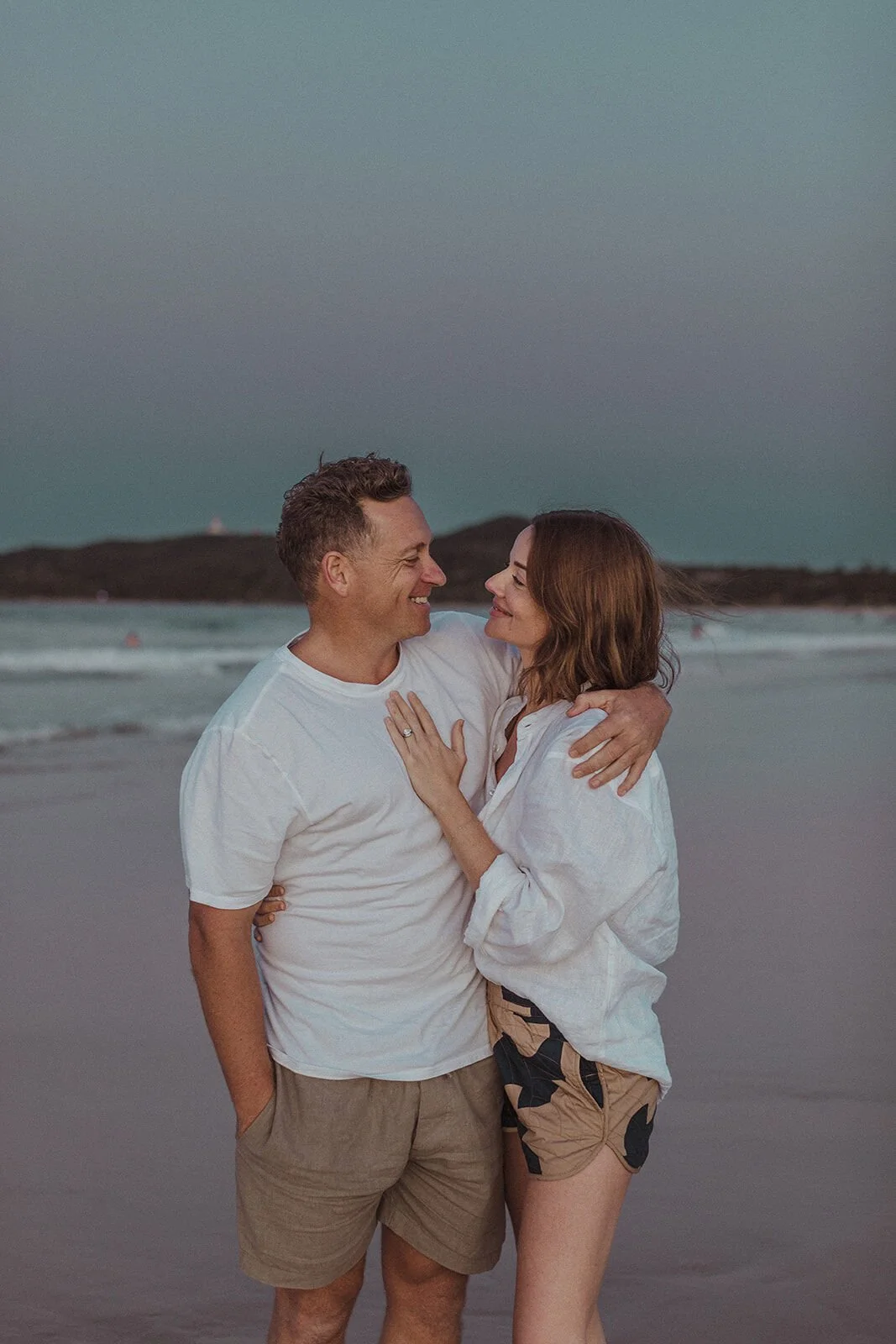 Couple smiling at each other on the beach at dusk, Jamie and Jessica's beach marriage proposal at Belongil Beach, Byron Bay