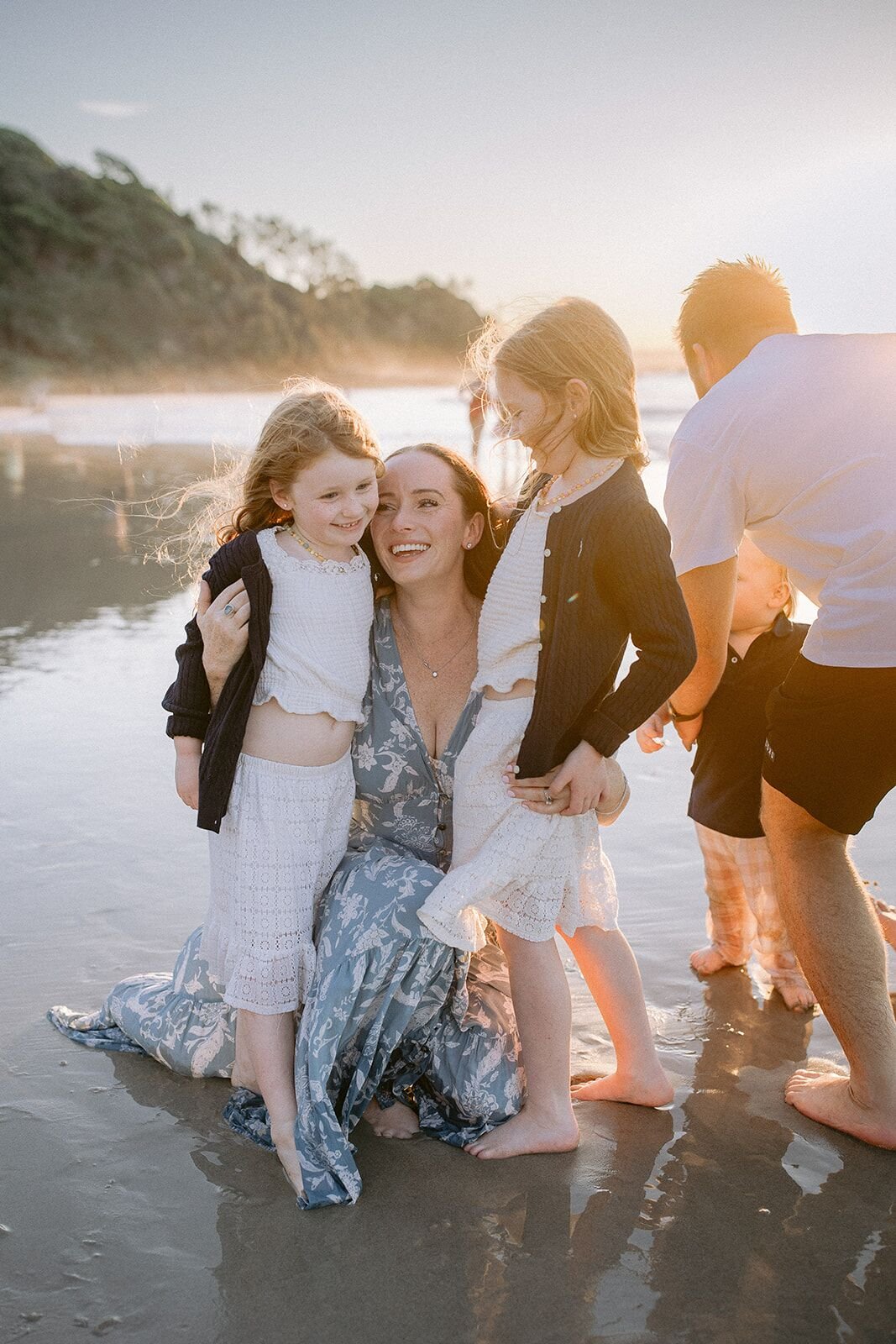 Mum kneeling on the wet sand laughing with her two daughters, dad and the green headland behind