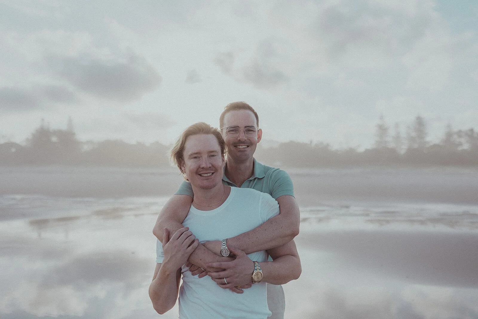 Couple embracing from behind and smiling at the camera on the beach, Byron and Luke's marriage proposal at Cabarita Beach, Northern Rivers