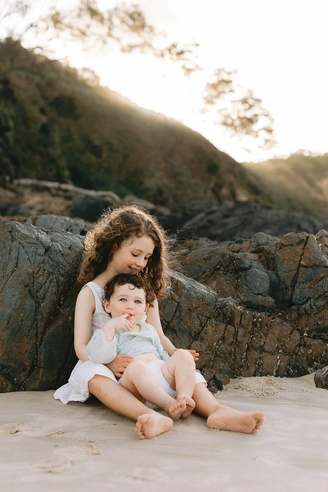 Brother and sister sitting together on the sand in front of the headland at sunset