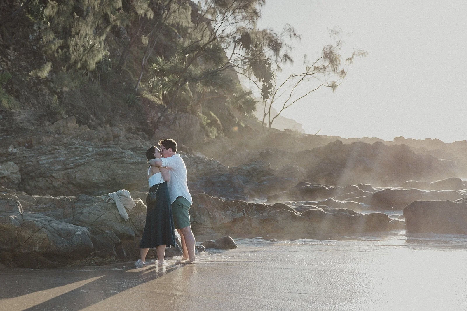 Couple embracing on the rocks in misty golden light, John and Emma's sunset marriage proposal at Wategos Beach, Byron Bay