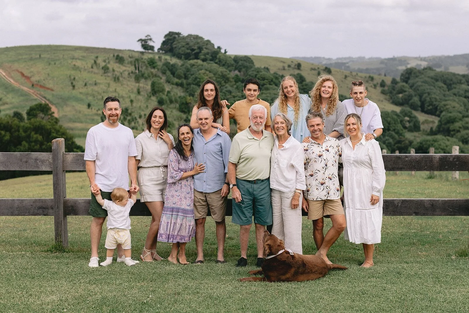 Extended family of three generations standing at a timber fence with a brown dog, green hills behind