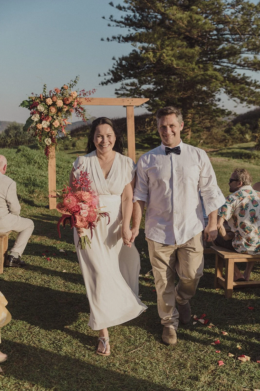 Couple walking down the aisle past guests beside a floral arch, Maritess and Mark's elopement at Lennox Head