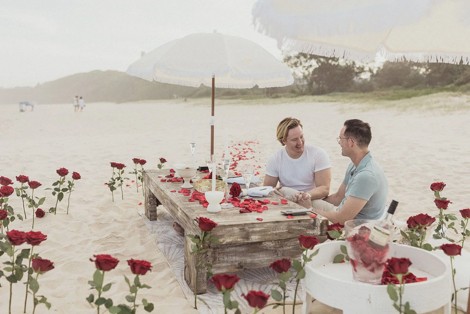Couple laughing together at the picnic table among red roses, Byron and Luke's marriage proposal at Cabarita Beach, Northern Rivers