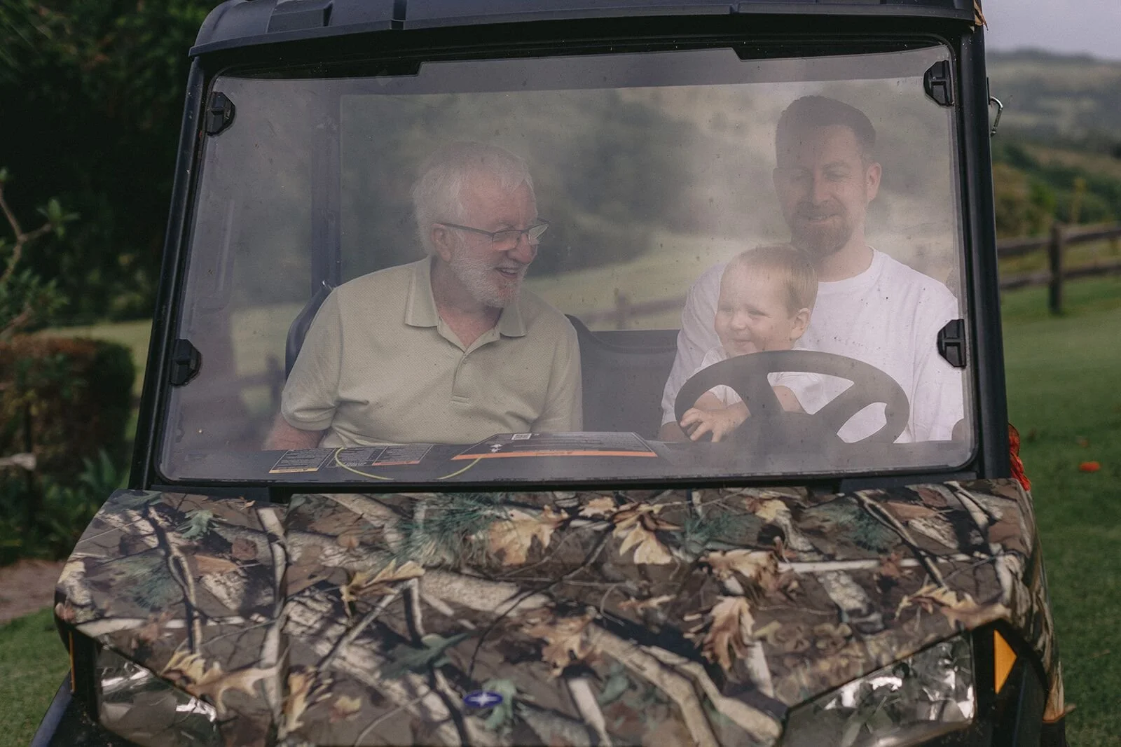Grandfather and baby grandson laughing behind the windscreen of a camouflage farm buggy