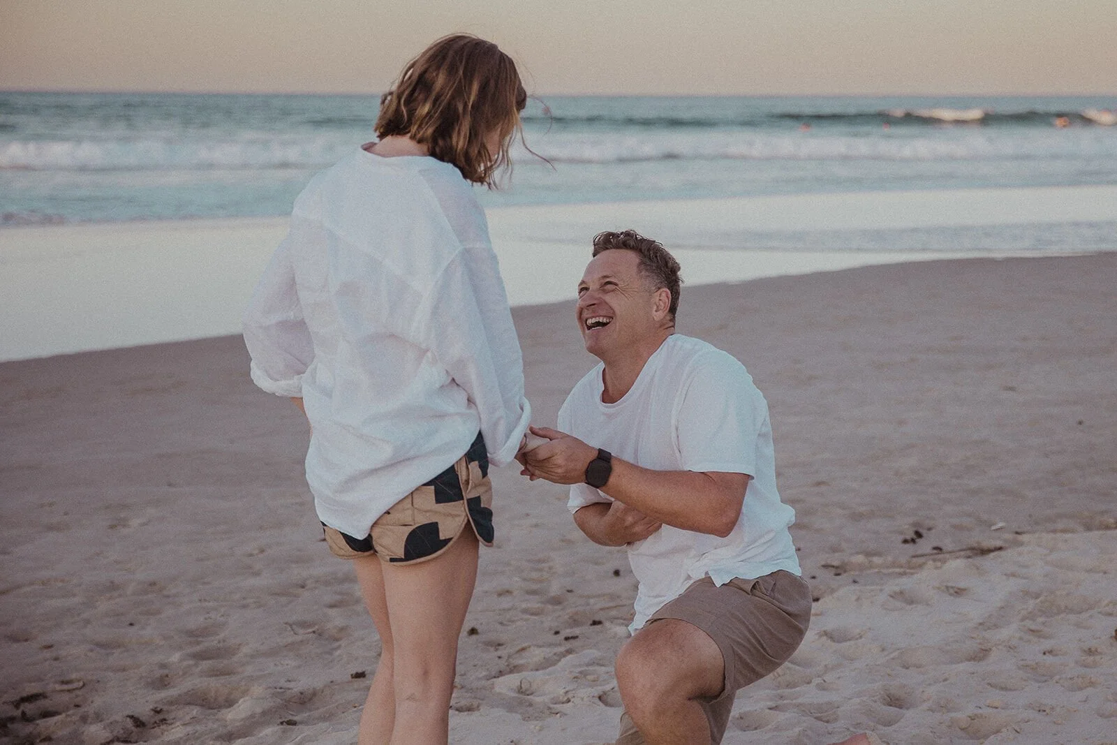 Man laughing on one knee as he holds up the ring, Jamie and Jessica's beach marriage proposal at Belongil Beach, Byron Bay