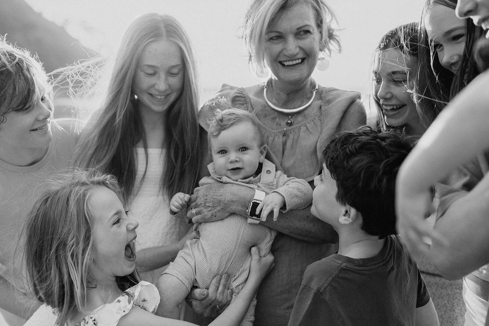 Black and white close-up of a laughing family group huddled around a baby and a grandmother in a pearl necklace