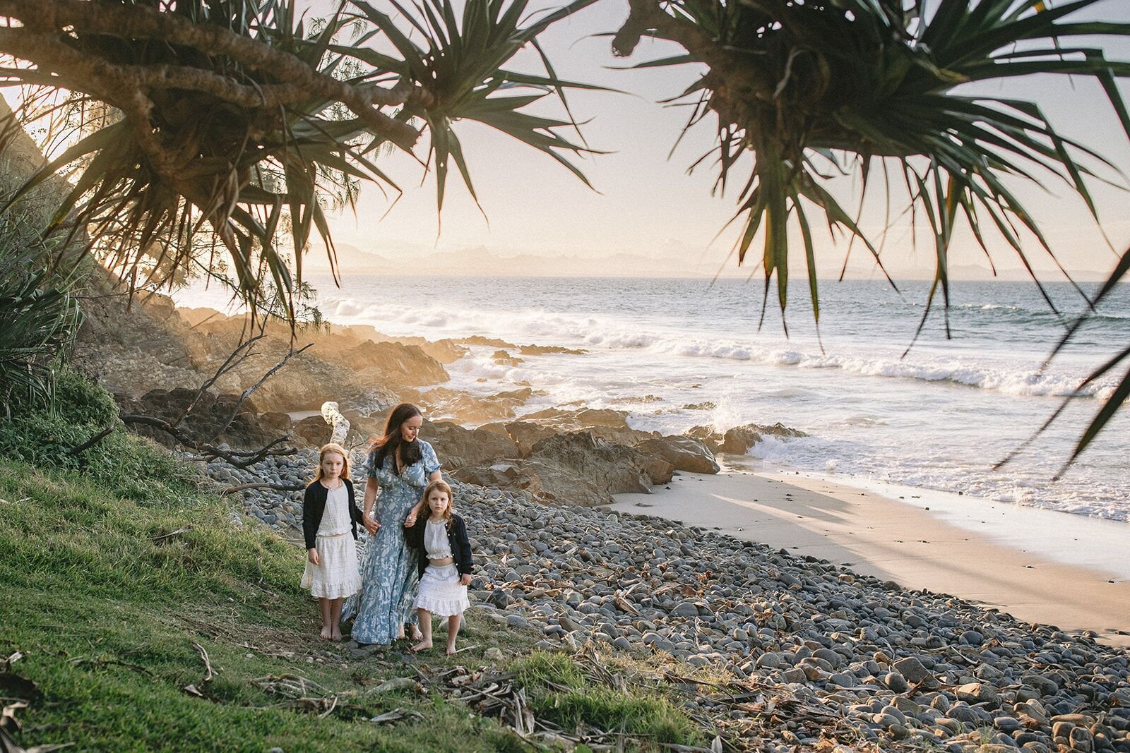 Family standing on the rocky shore at Wategos Beach under pandanus palms with the ocean behind