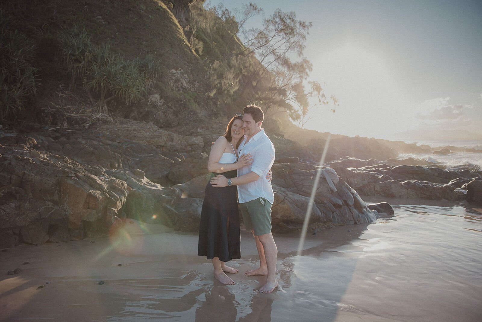 Couple embracing in the golden sun flare on the shore, John and Emma's sunset marriage proposal at Wategos Beach, Byron Bay