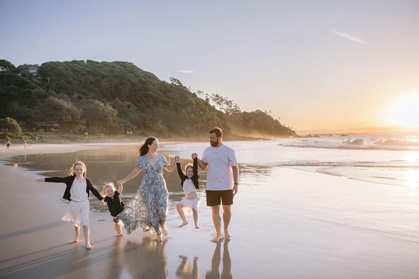 Family walking hand in hand along the wet sand at Wategos Beach with the headland and setting sun behind