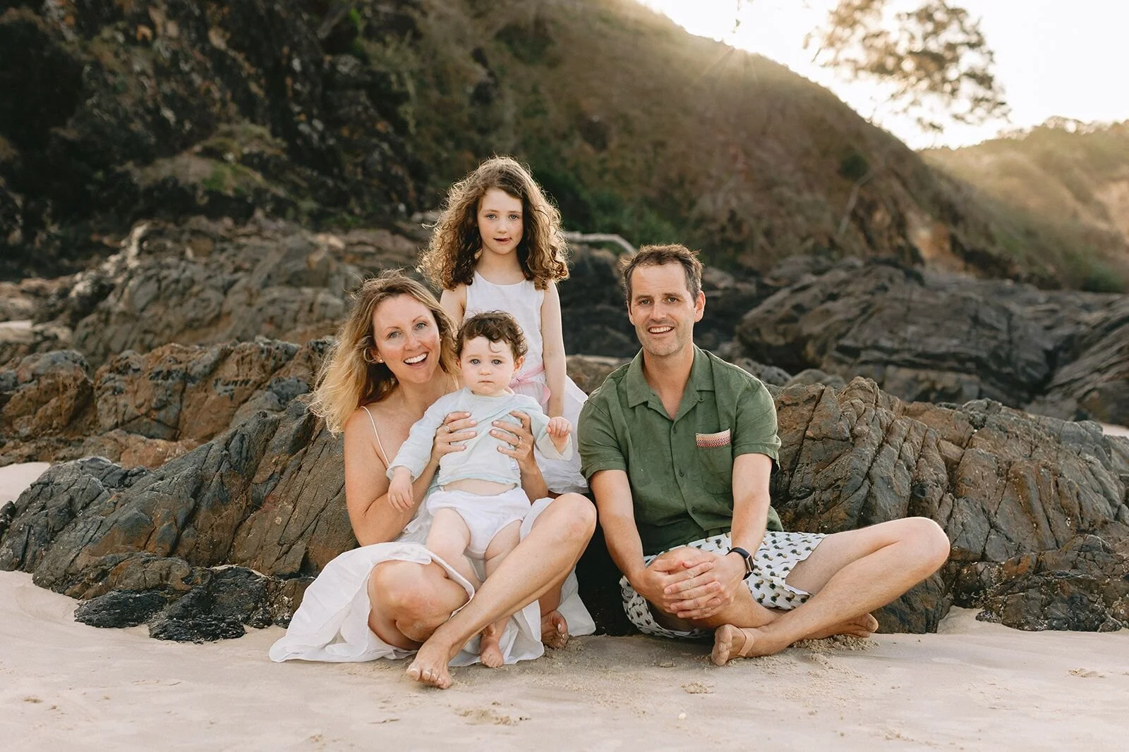 Family sitting together on the sand in front of the rocks, mum holding the toddler and dad smiling beside them