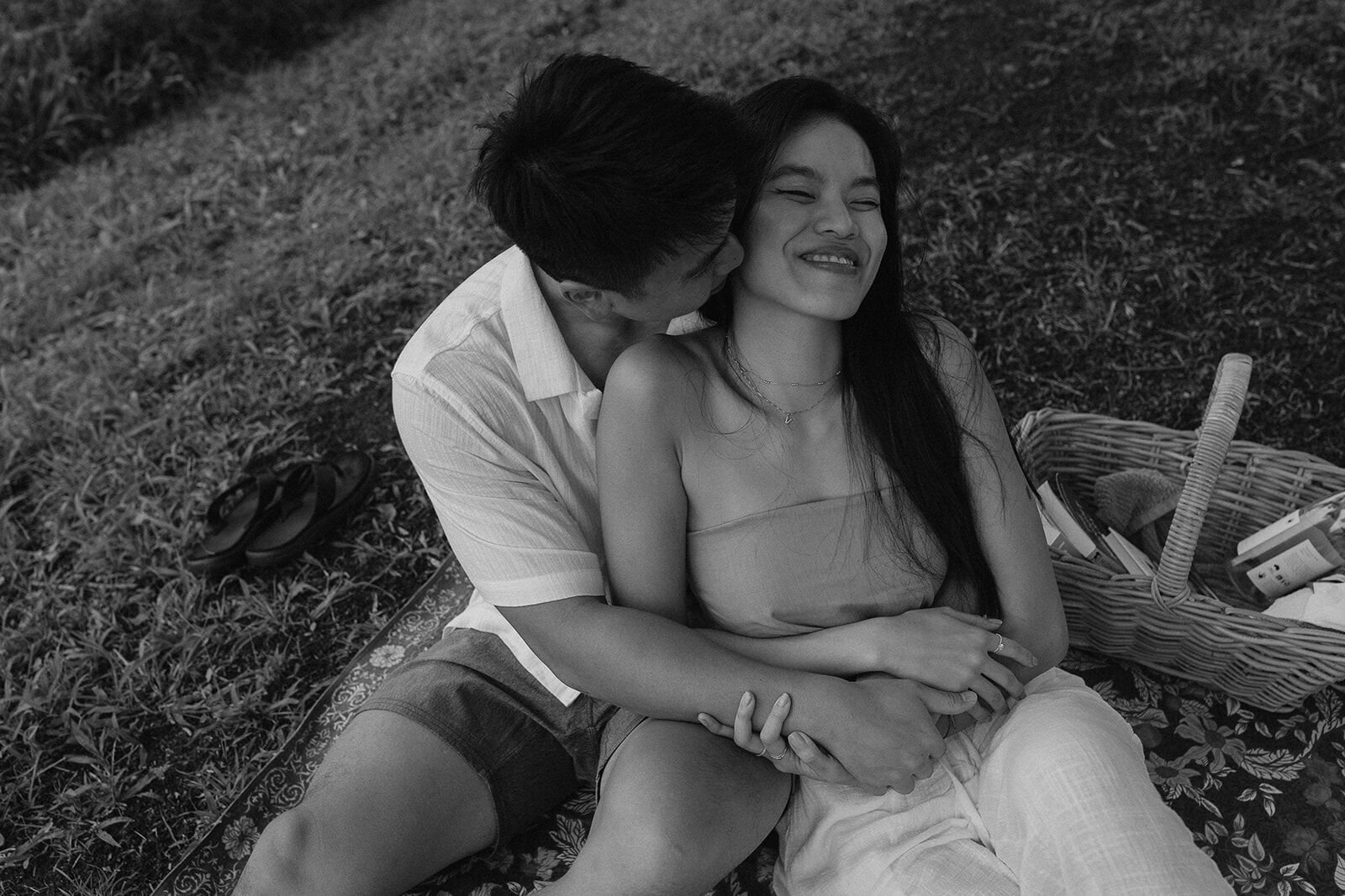 Black and white of the couple laughing and cuddling beside the picnic basket, Darien and Jacqui's picnic marriage proposal at Heartwood Farm, Byron Bay hinterland