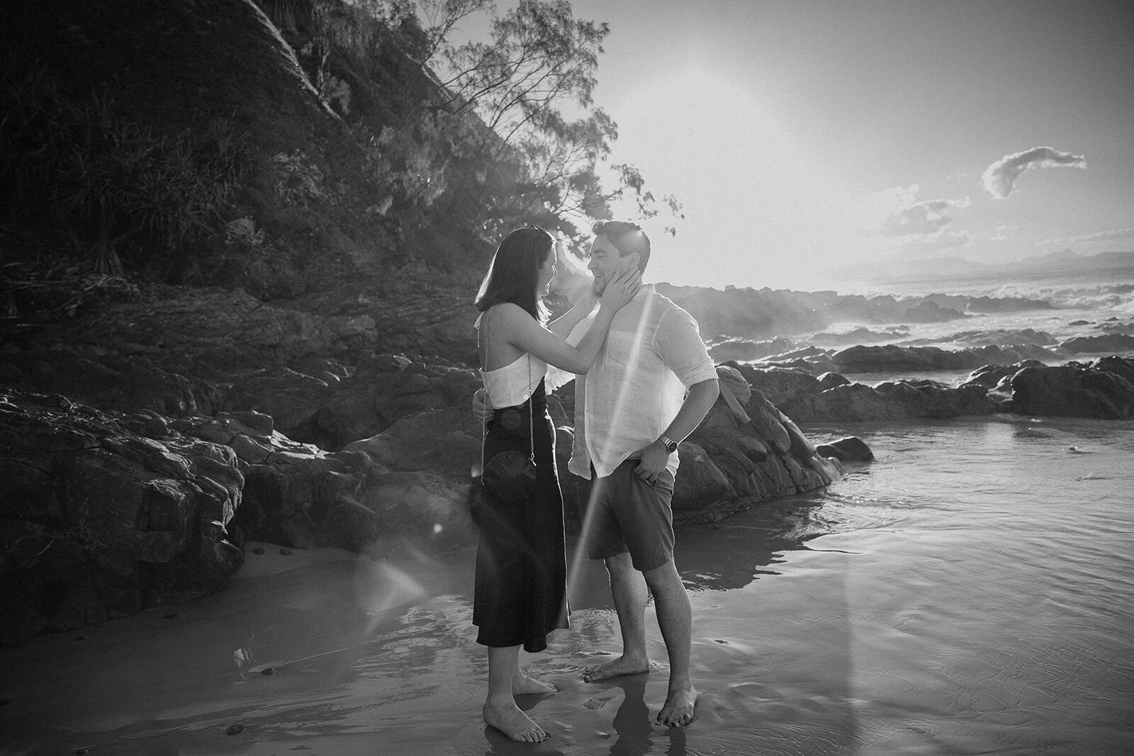 Black and white of the woman touching her fiancé's face with the sun behind them, John and Emma's sunset marriage proposal at Wategos Beach, Byron Bay