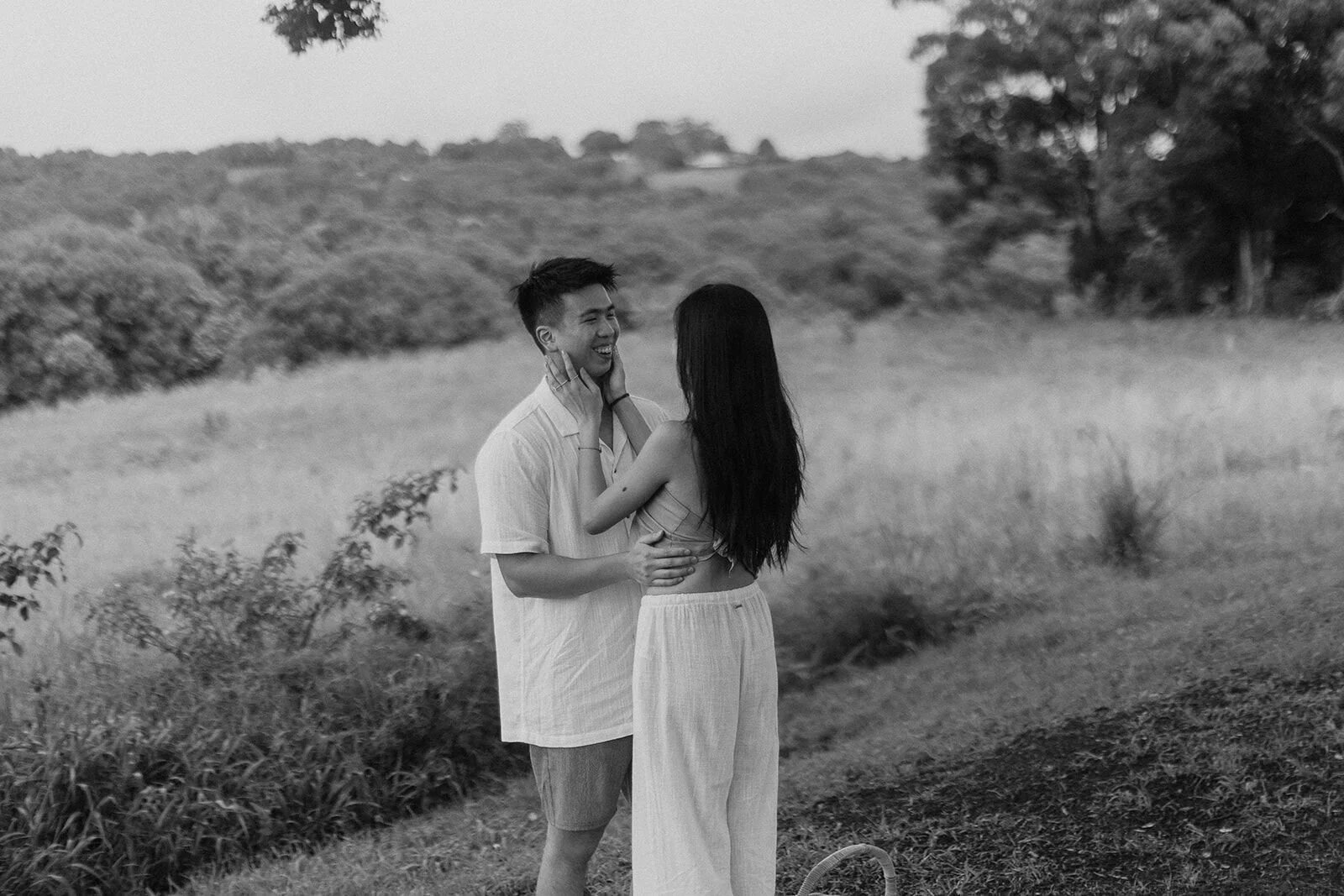 Black and white of the woman touching her partner's face in the paddock, Darien and Jacqui's picnic marriage proposal at Heartwood Farm, Byron Bay hinterland