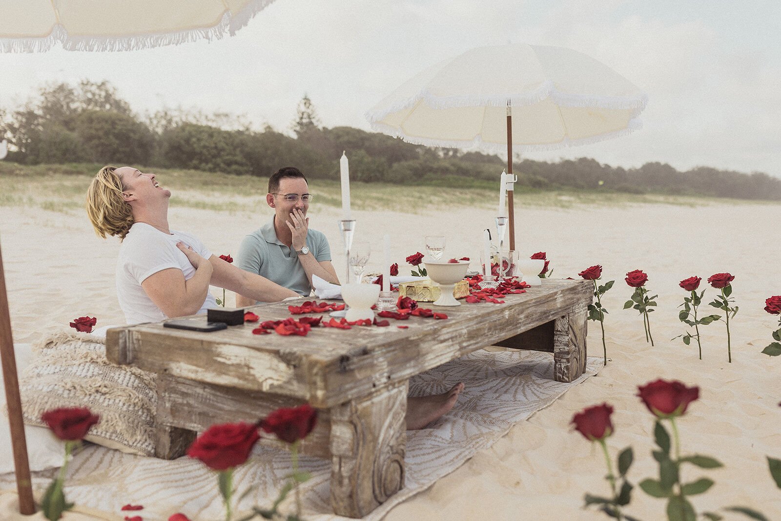 Couple laughing at the table on the sand with a ring box in front of them, Byron and Luke's marriage proposal at Cabarita Beach, Northern Rivers