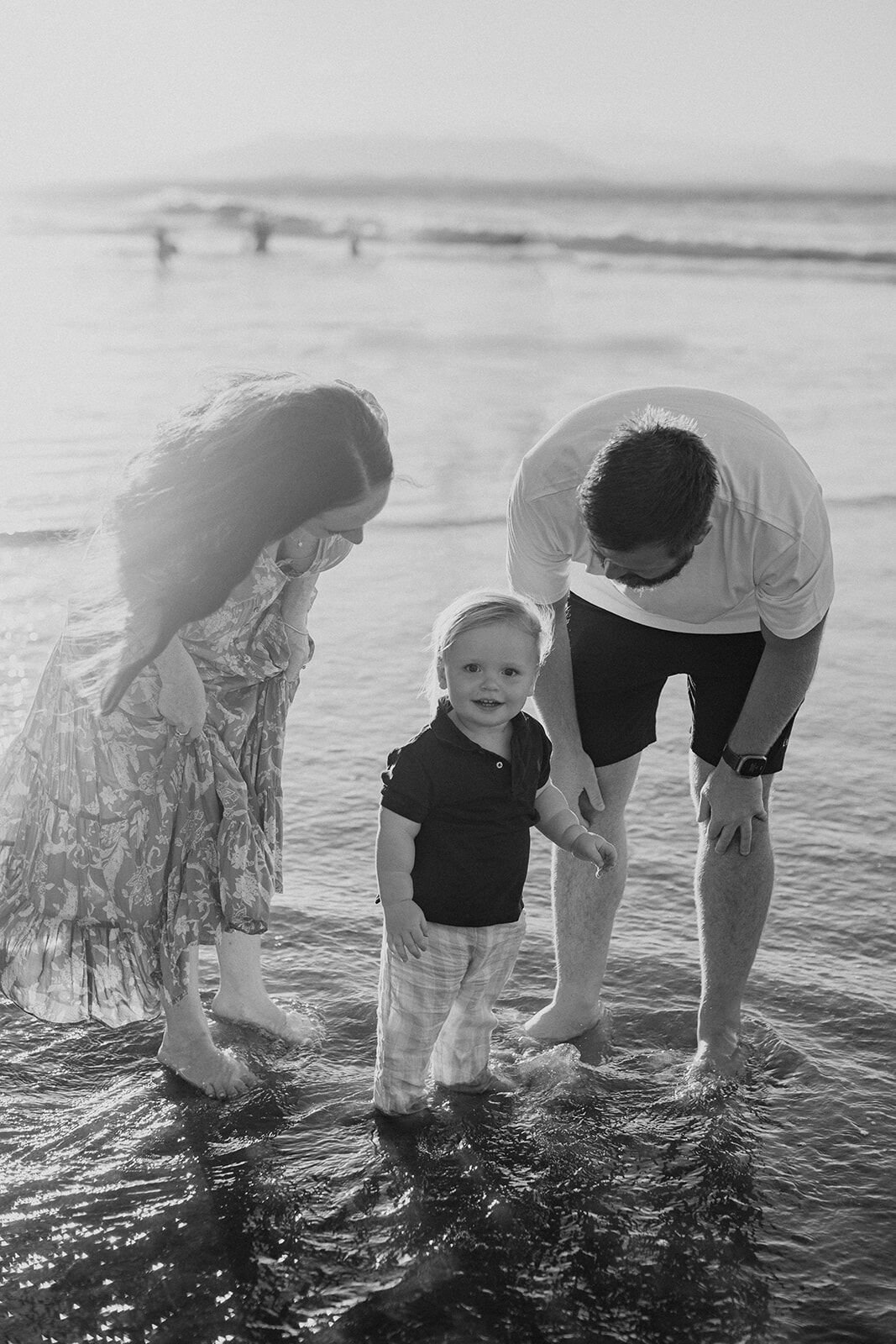 Black and white photo of mum and dad with their toddler paddling in the shallows
