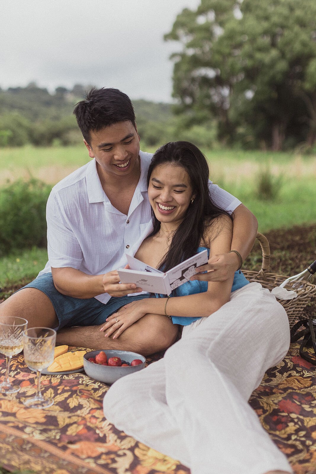 Couple laughing together over a small book on the picnic rug, Darien and Jacqui's picnic marriage proposal at Heartwood Farm, Byron Bay hinterland