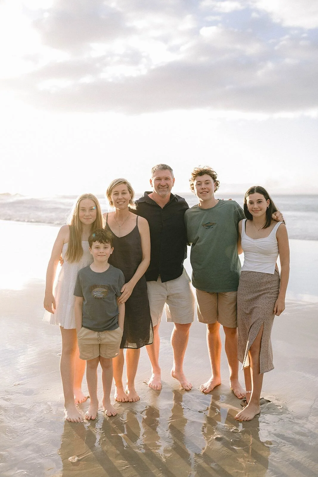Parents and four teenage and younger kids standing in a row on the wet sand at sunset