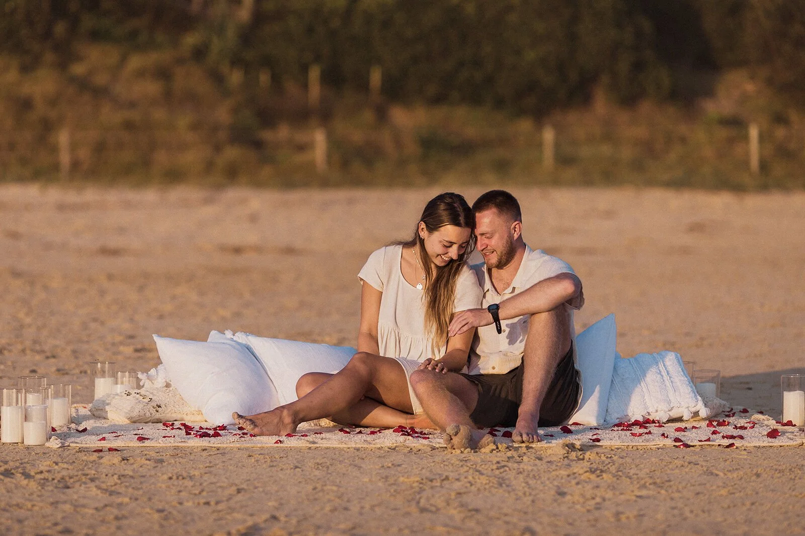 Couple sitting together on the pillows and smiling at the ring, Charlie and Hannah's sunrise marriage proposal at Broken Head Beach, Byron Bay