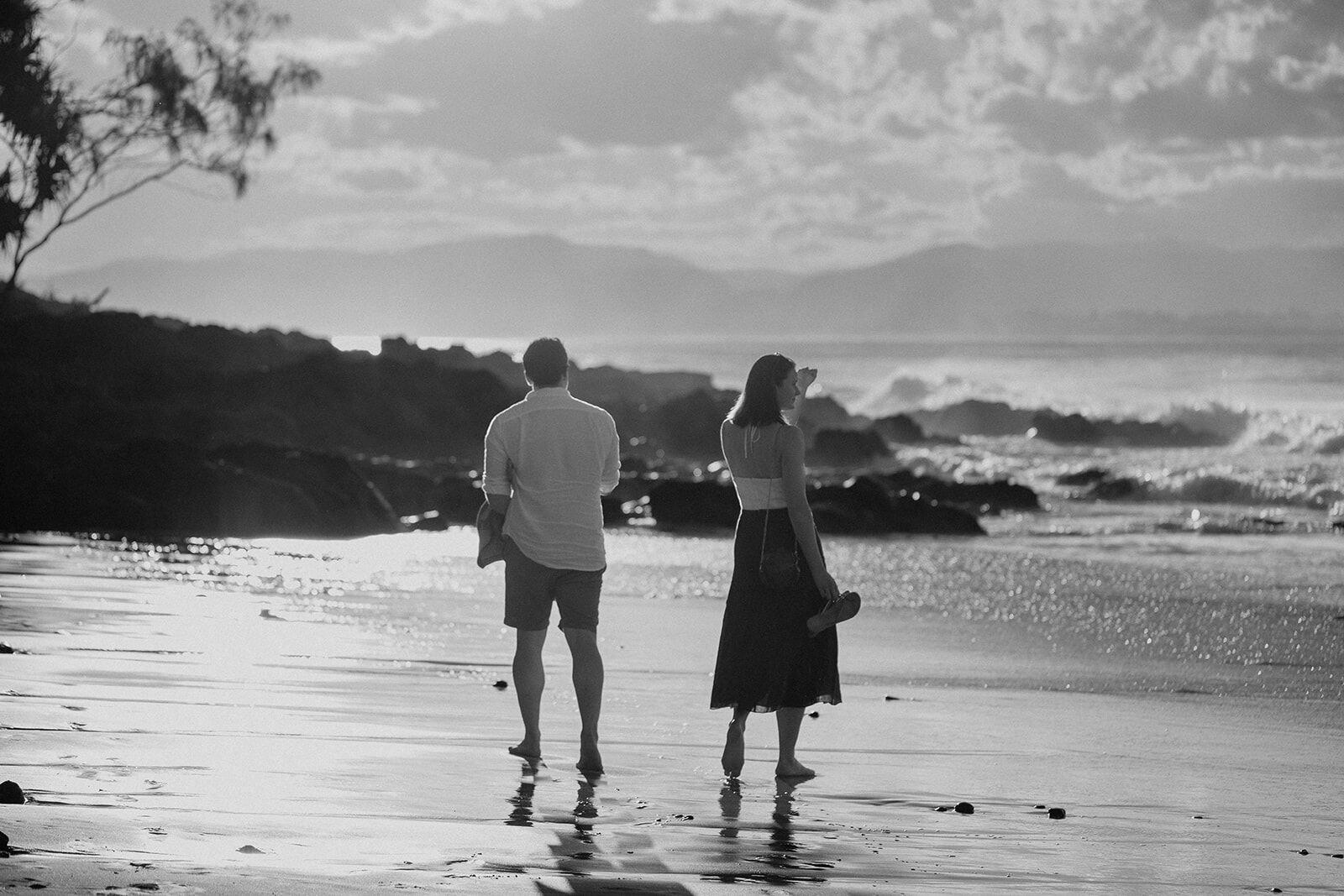 Black and white of the couple walking along the shoreline toward the rocks at Wategos Beach, John and Emma's sunset marriage proposal at Wategos Beach, Byron Bay