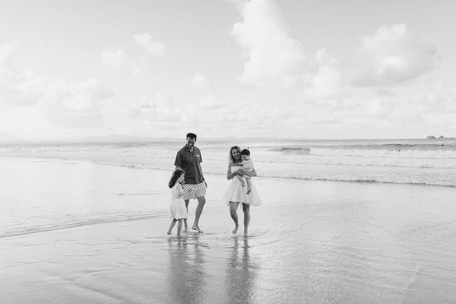 Black and white wide shot of a family wading through shallow water on a quiet beach