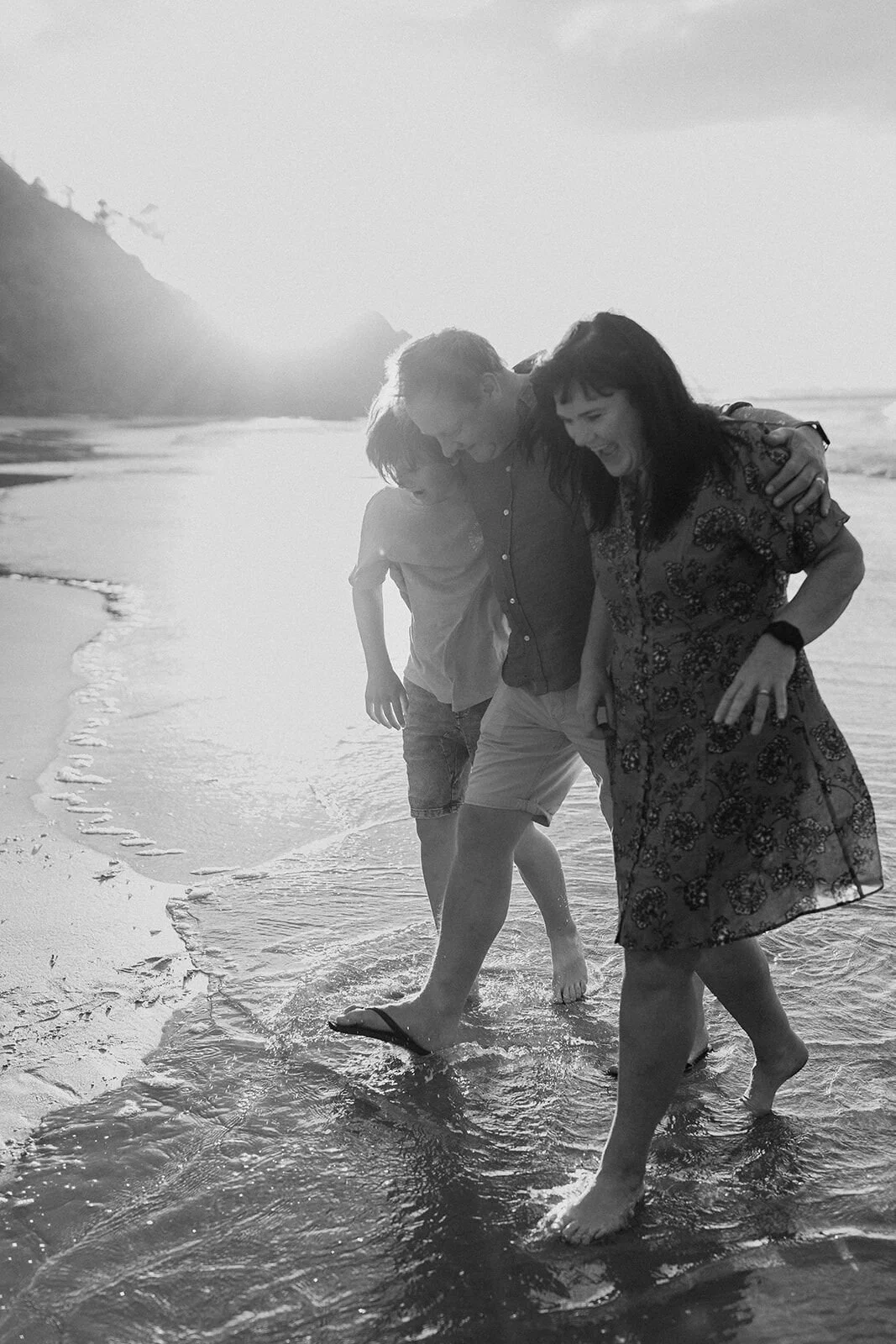 Black and white photo of a mum and her son walking through the shallows with the sun behind the headland
