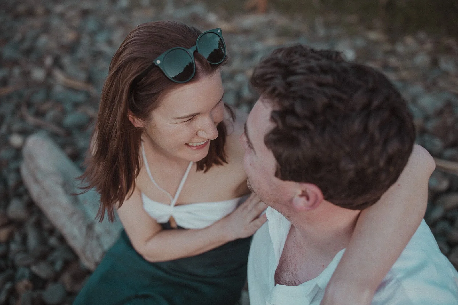 Couple smiling at each other on the pebbly beach, her sunglasses on her head, John and Emma's sunset marriage proposal at Wategos Beach, Byron Bay