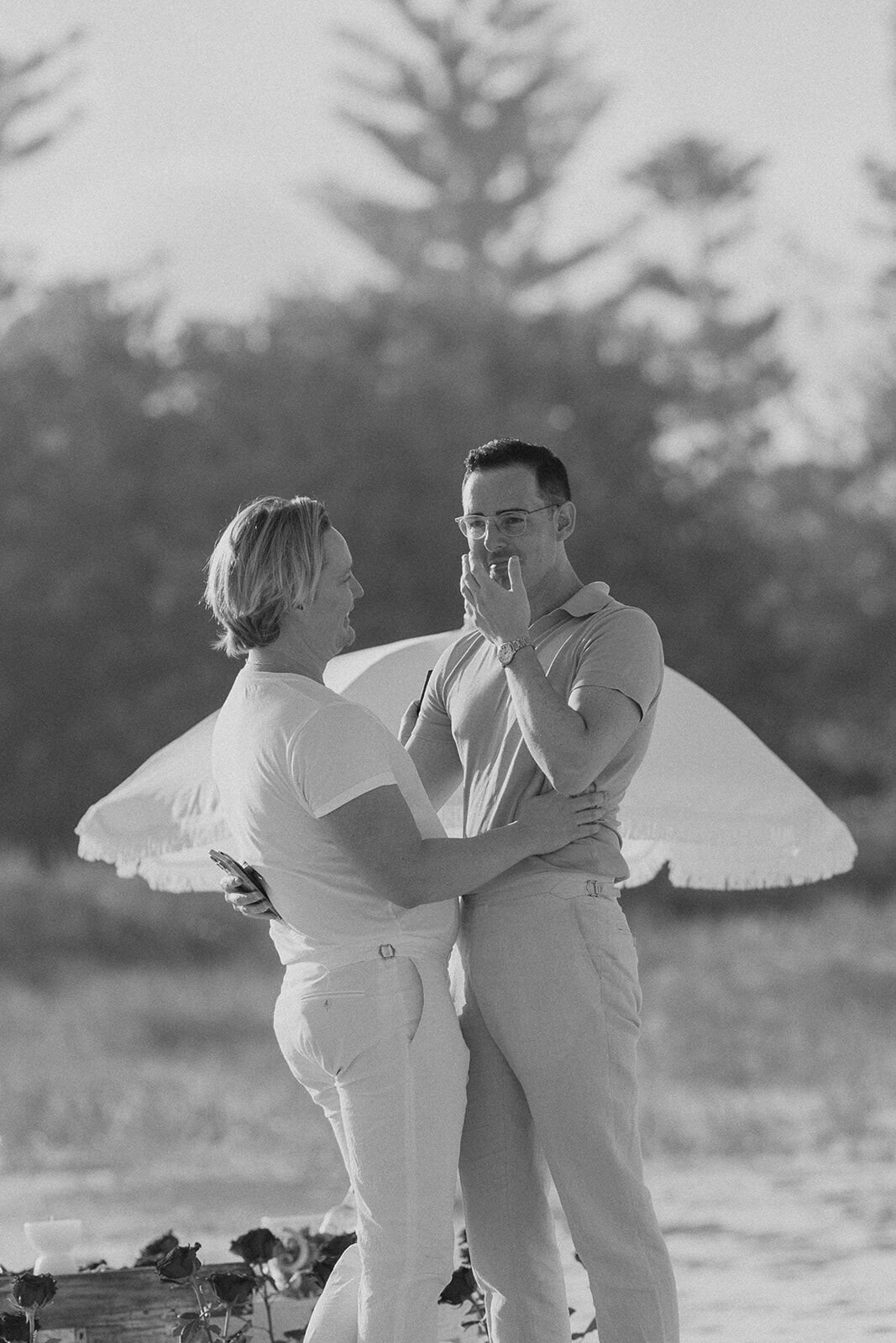 Black and white of the couple embracing with one covering his mouth in surprise, Byron and Luke's marriage proposal at Cabarita Beach, Northern Rivers