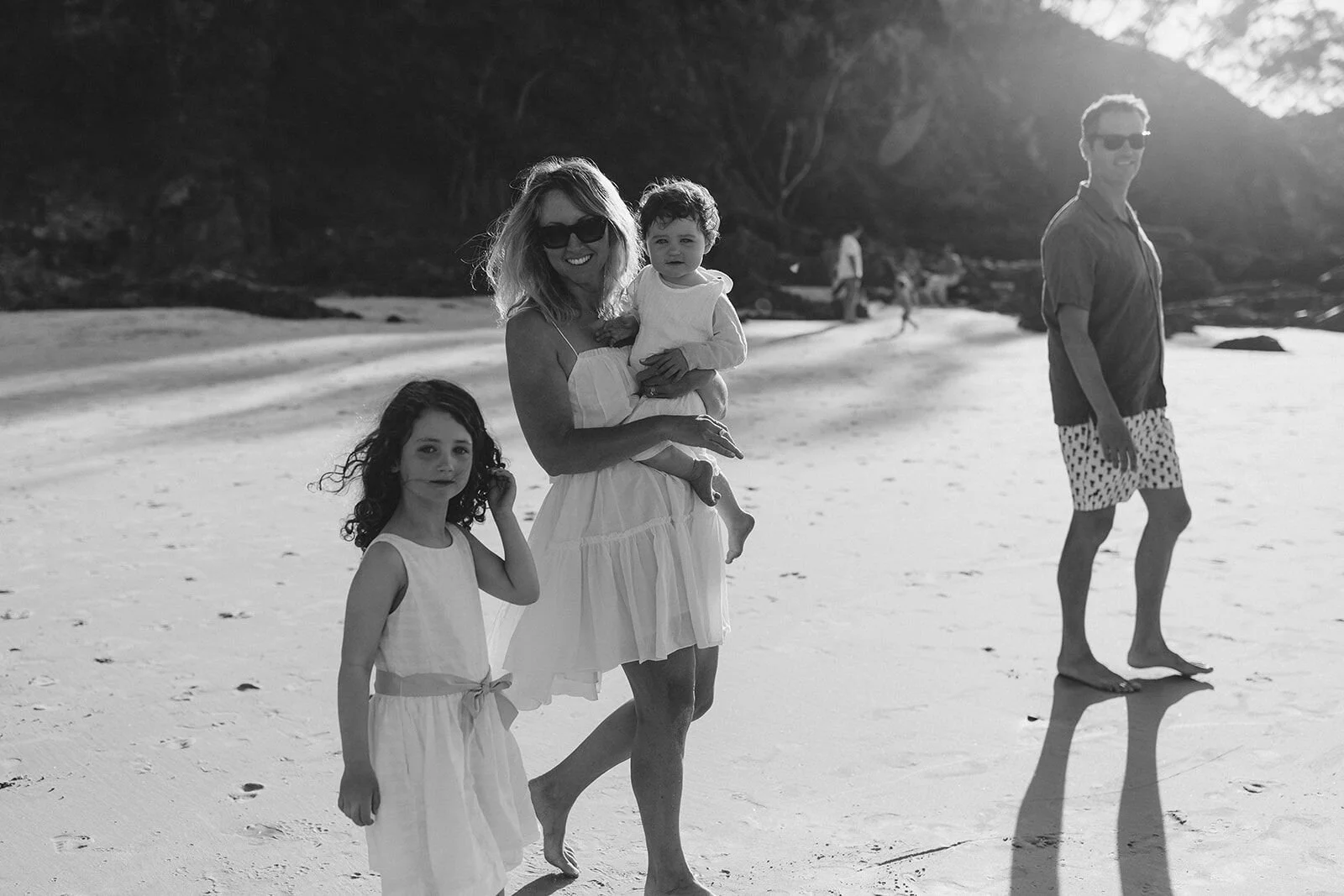 Black and white photo of a family walking along Wategos Beach, Byron Bay, with mum carrying their toddler