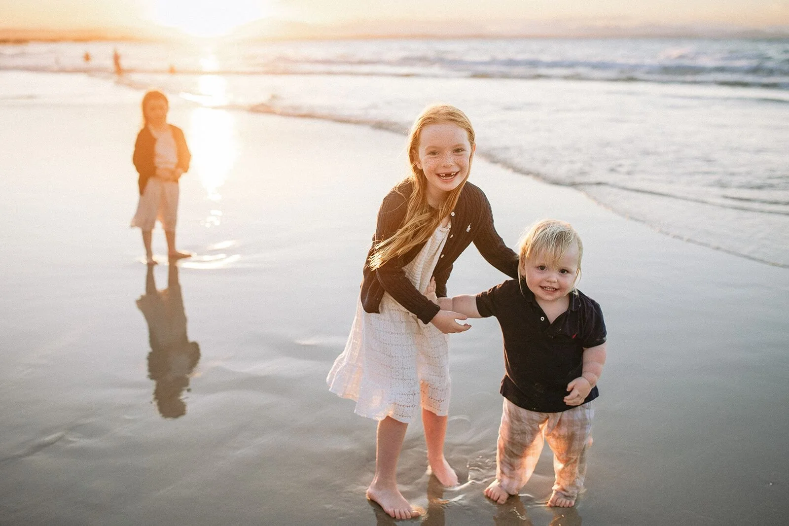A girl and her toddler brother holding hands at the water's edge, their sister in the distance