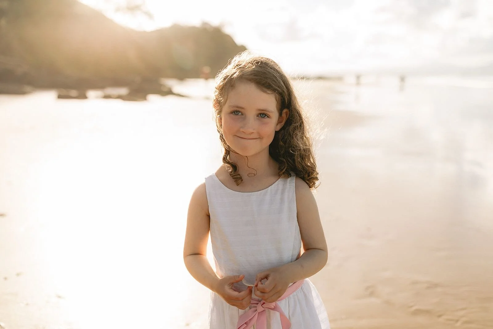 Girl in a white dress smiling softly at the camera on the beach at sunset