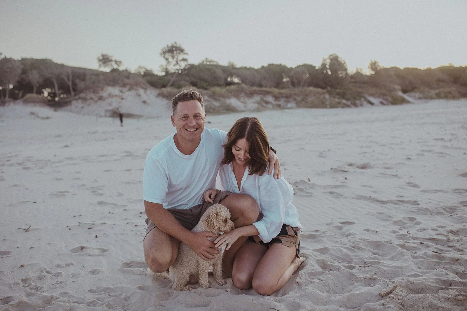 Couple kneeling on the sand and smiling with their dog, Jamie and Jessica's beach marriage proposal at Belongil Beach, Byron Bay