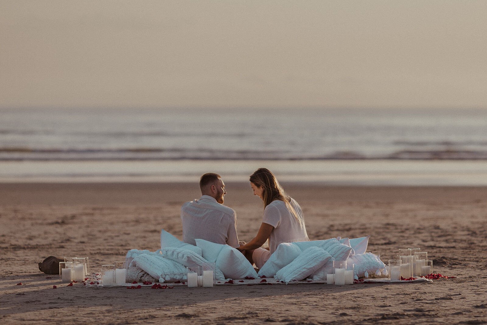 Couple sitting on the pillows facing the ocean at sunrise, Charlie and Hannah's sunrise marriage proposal at Broken Head Beach, Byron Bay