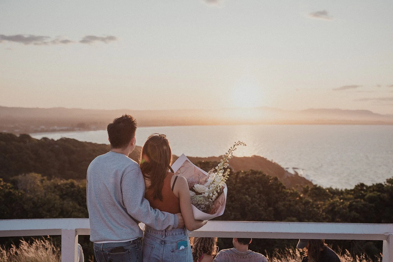 Couple from behind with the bouquet, watching the sun go down over the bay, Douglas and Ying's sunset marriage proposal at Cape Byron Lighthouse, Byron Bay