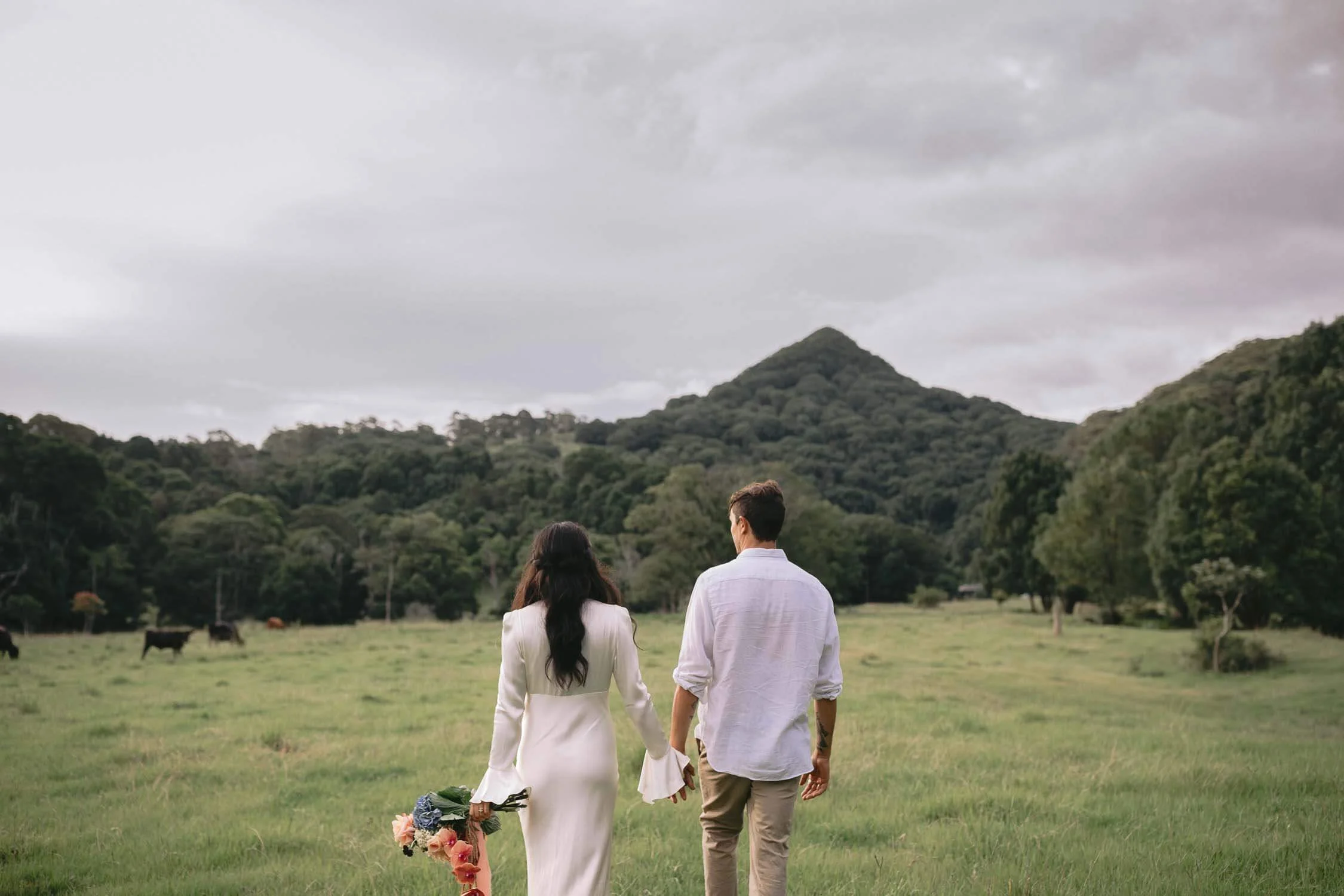 Eloping couple walking across a paddock towards Mount Chincogan in the Byron hinterland
