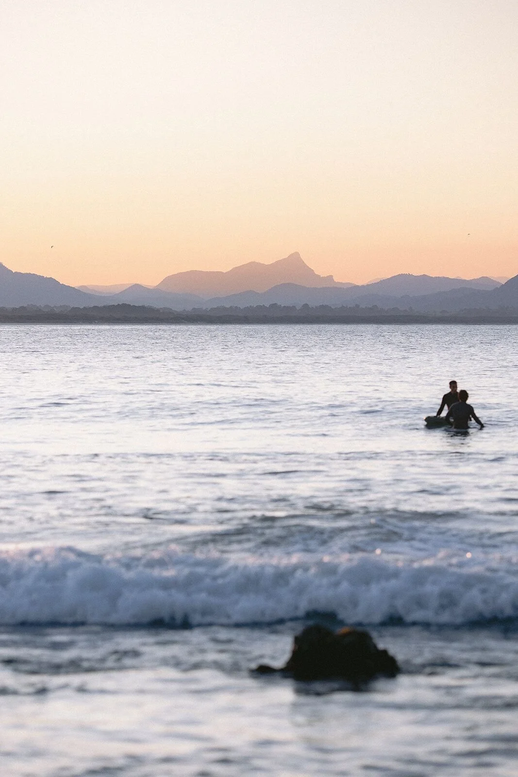 Surfer paddling out beside a rock at dusk with Wollumbin on the horizon