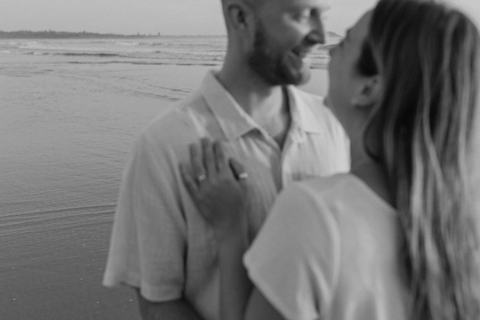 Black and white close up of the couple smiling with her ringed hand on his chest, Charlie and Hannah's sunrise marriage proposal at Broken Head Beach, Byron Bay
