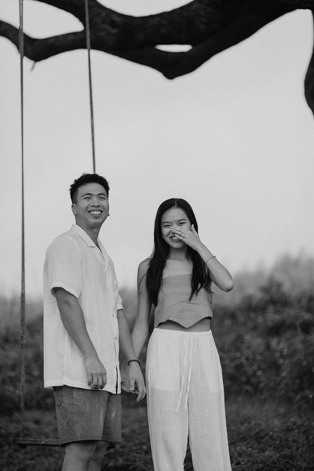Black and white of the couple holding hands, the woman laughing and covering her face, Darien and Jacqui's picnic marriage proposal at Heartwood Farm, Byron Bay hinterland