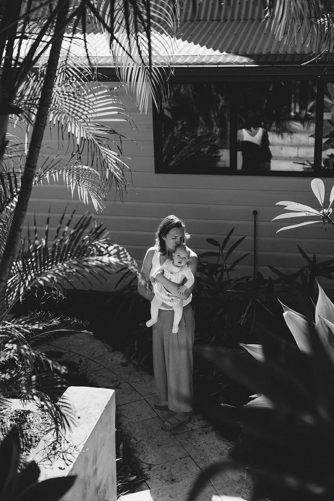 Black and white photo of mum carrying her baby on a garden path beside the house