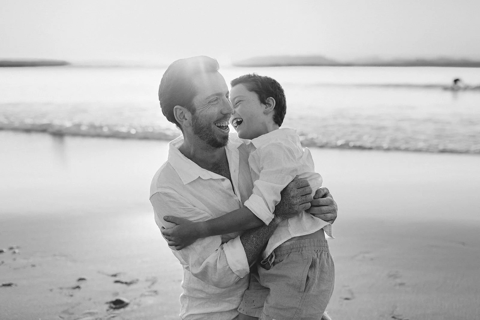 Black and white photo of dad and son laughing together in a bear hug at the shoreline