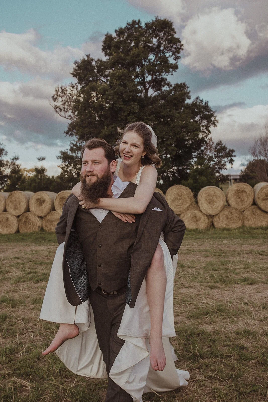 Groom carrying the bride piggyback beside hay bales, Sam and Tom's elopement at Highfield Farm, Northern Rivers