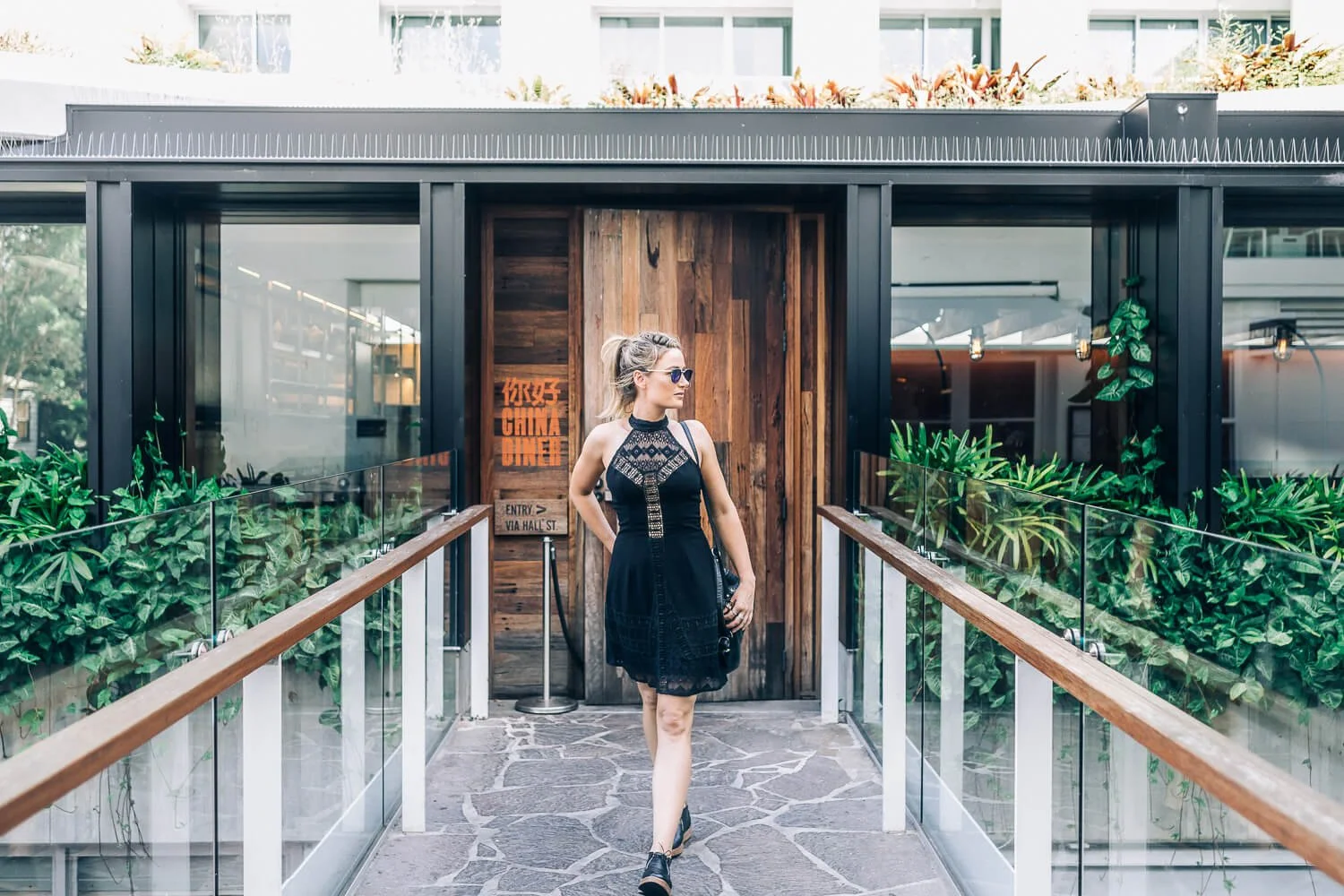Woman in a black dress walking across a bridge from a timber-doored venue, Bondi lifestyle shoot