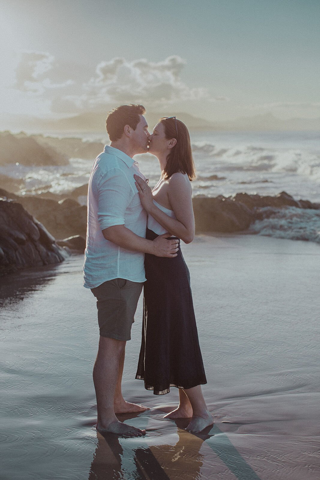 Couple kissing in the shallows with waves breaking behind them, John and Emma's sunset marriage proposal at Wategos Beach, Byron Bay
