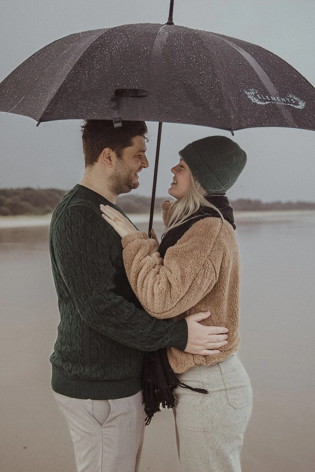 Couple smiling at each other under the umbrella on the wet sand, Raphael and Vany's rainy day marriage proposal at Broken Head Beach, Byron Bay