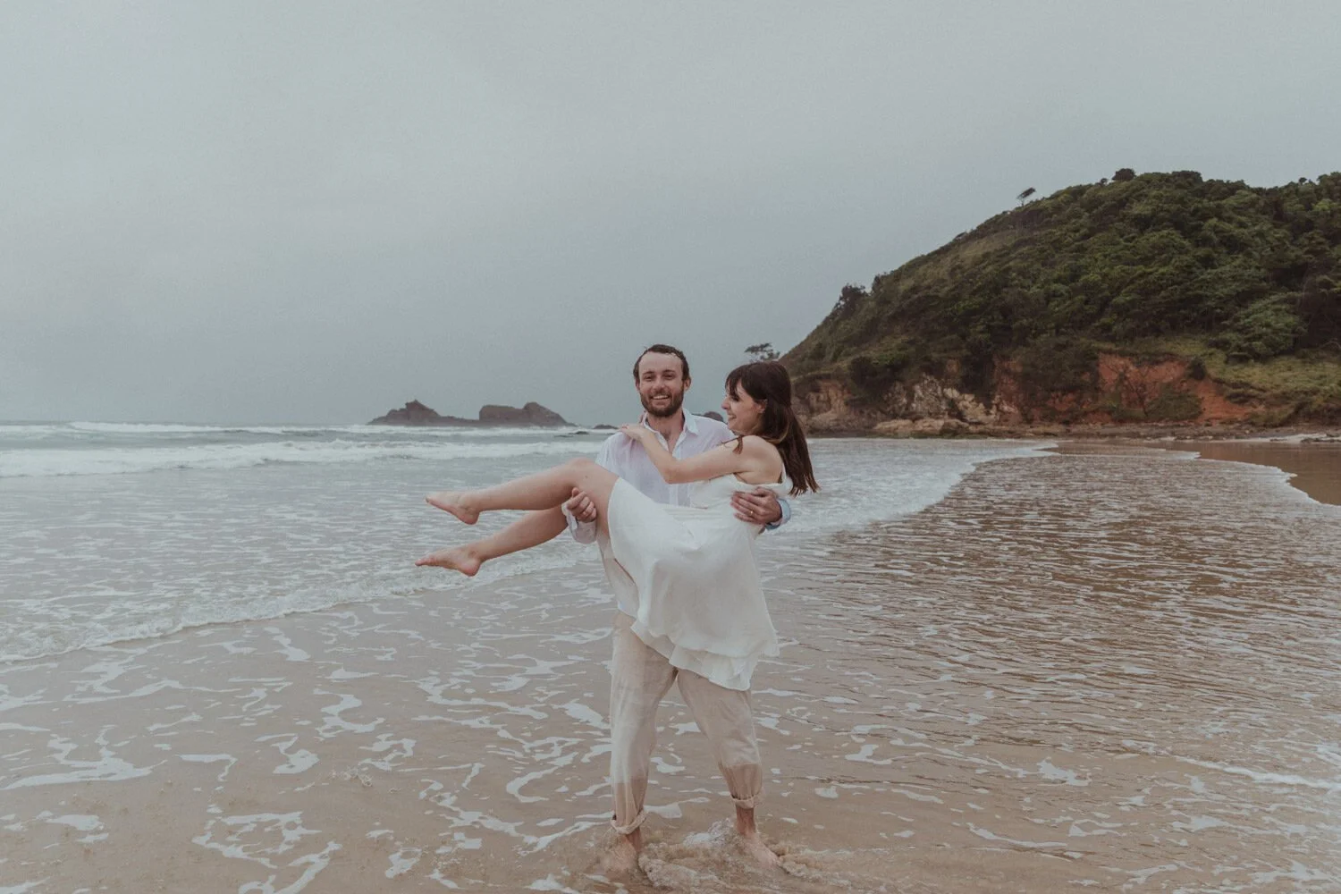 Groom carrying the bride through the surf, Yoanne and Elisa's rainy elopement at Broken Head, Byron Bay