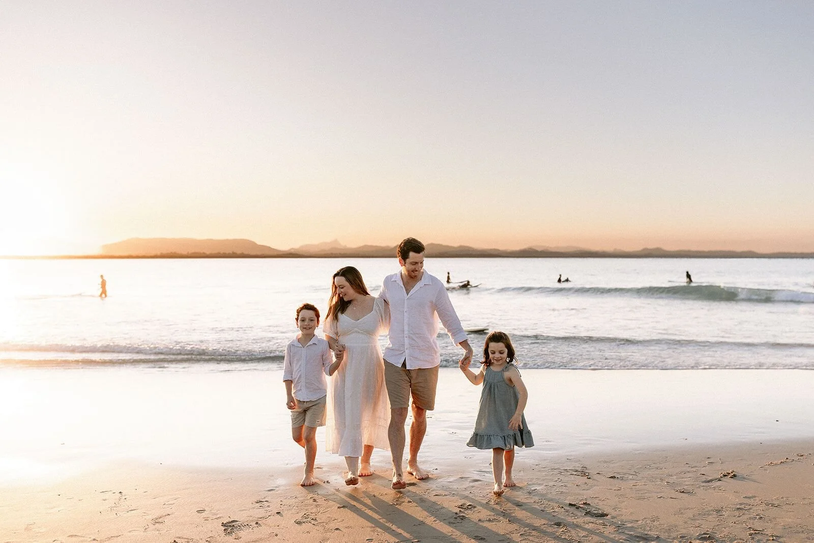 Family walking toward the camera along the sand at sunset, hand in hand