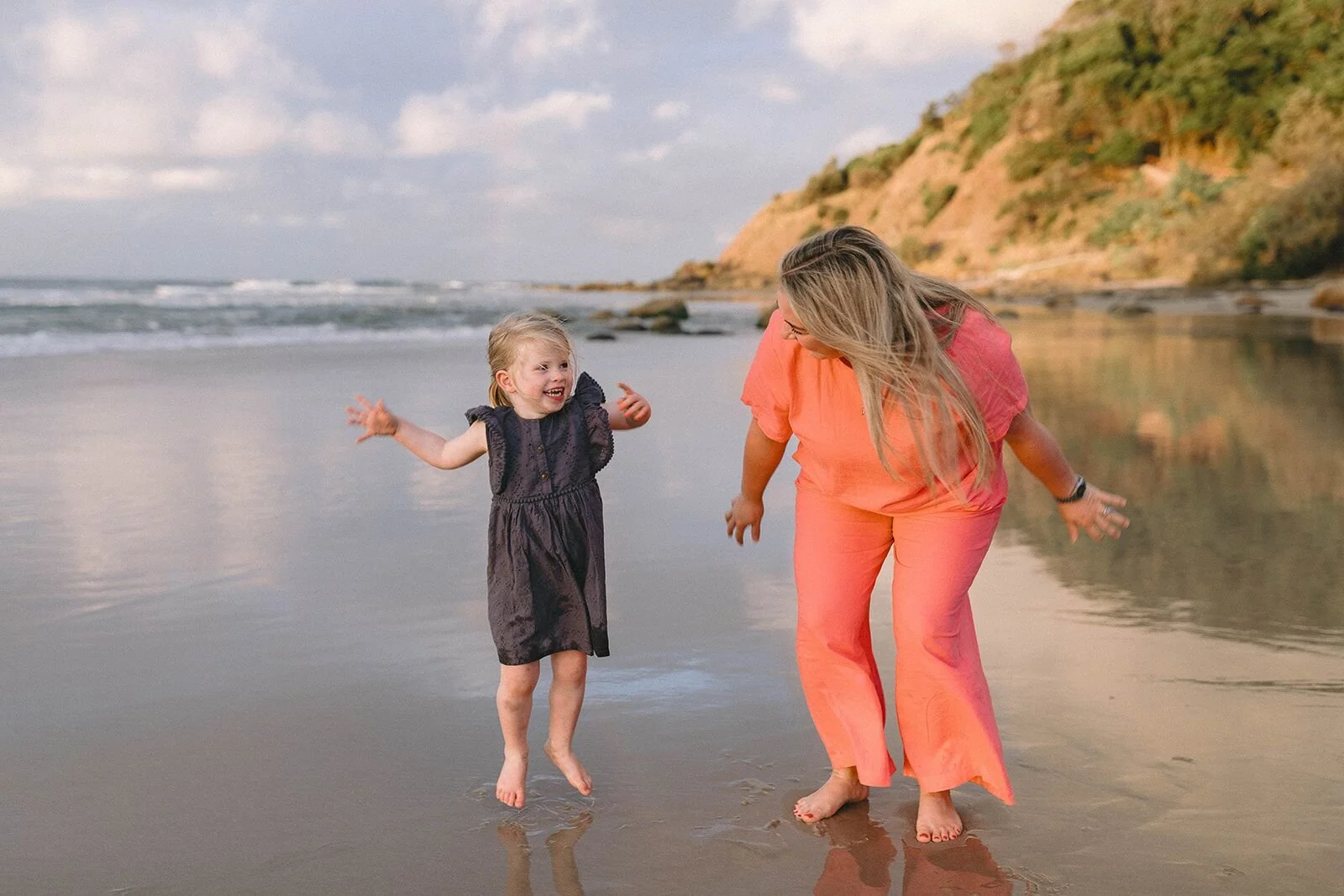 Toddler in a navy dress dancing on the sand while her aunt in a coral outfit plays with her